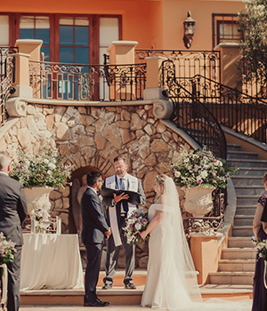 Outdoor wedding ceremony with a couple standing before an officiant, with stairs and a balcony in the background.