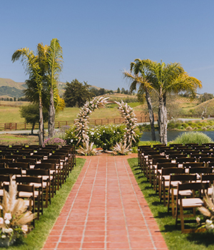 Outdoor wedding ceremony setup with chairs and a floral archway on a sunny day.