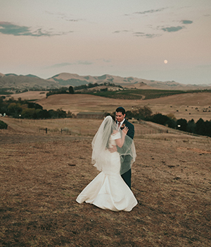 A couple in wedding attire embracing in a field at dusk, with rolling hills in the background and a pale sky above.