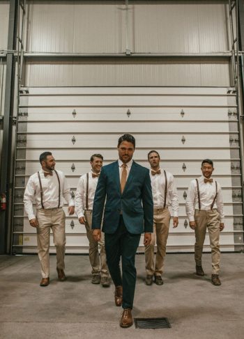 A man in a blue suit leads a group of men in white shirts and suspenders walking inside a warehouse.