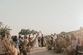 Outdoor wedding ceremony with guests seated on either side of an aisle leading to a triangular arch where a couple stands facing each other.