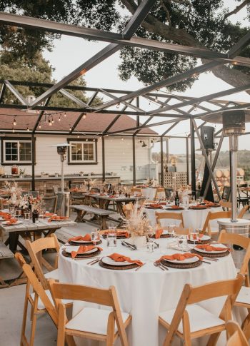 Outdoor dining setup with wooden tables and chairs under a pergola, featuring elegant table settings and ambient lighting.