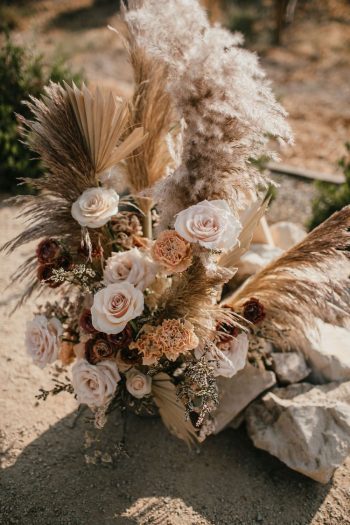 A rustic bouquet with white roses, dried grasses, and pampas grass set upon rocks.