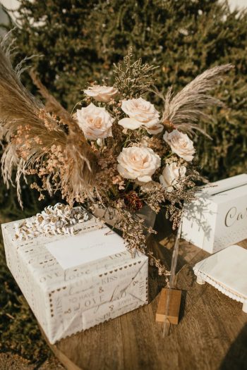 A wedding bouquet of cream roses and pampas grass next to a gift box on a wooden table.