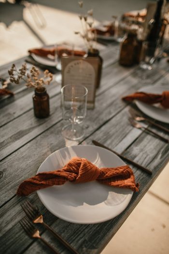 Rustic table setting with an empty white plate, an orange napkin, and copper cutlery, arranged for an outdoor event.