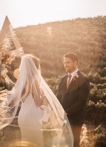 A bride and groom smiling at each other during an outdoor wedding ceremony with a geometric arch and natural backdrop.