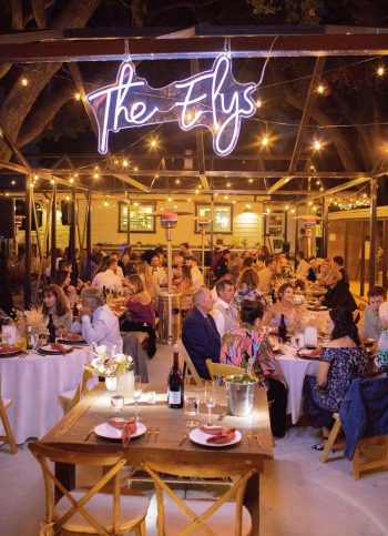 Elegant evening event at an outdoor venue with guests seated at dinner tables under a neon sign.