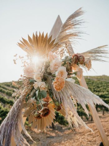 A sunlit floral arrangement with pampas grass and golden leaves against a vineyard backdrop.