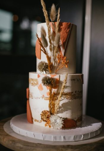 Three-tiered cake with abstract terracotta and gold accents, adorned with dried botanicals.