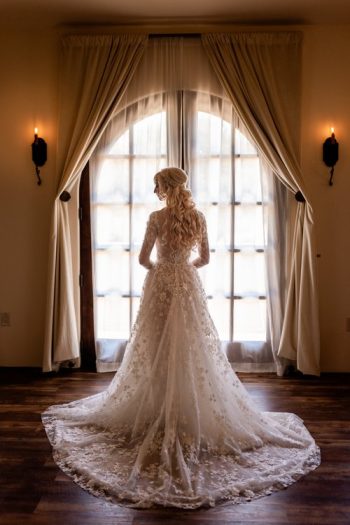 Bride in an elegant lace wedding dress standing in front of a window with sheer curtains.