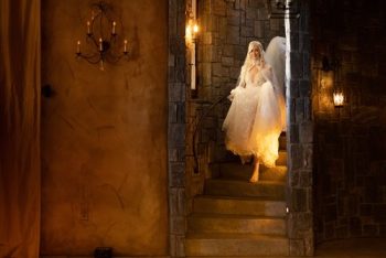 A bride in a white dress descending a stone staircase in a warmly lit, castle-like interior.
