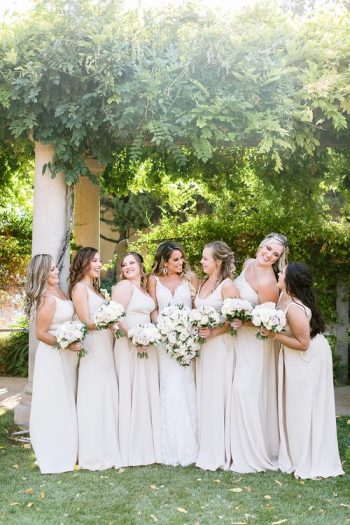 A bride with her bridesmaids, all smiling and holding bouquets, in a garden setting.
