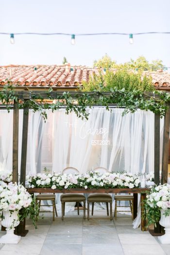 Elegant outdoor wedding reception area with a floral decorated table under a pergola, featuring drapery and string lights.