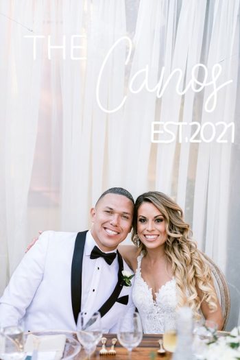 A smiling couple dressed in wedding attire seated at a table with the sign "the canos est.2021" displayed above them.