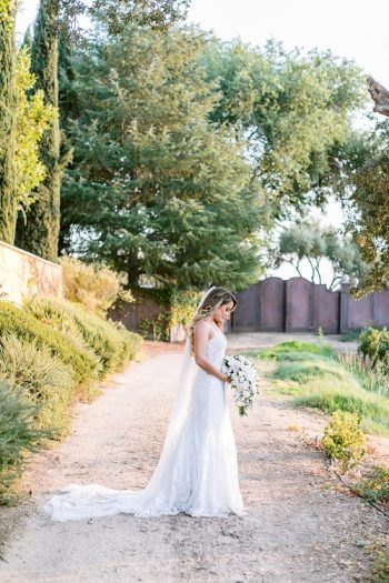 A bride holding a bouquet walks down a gravel path with greenery on the side.