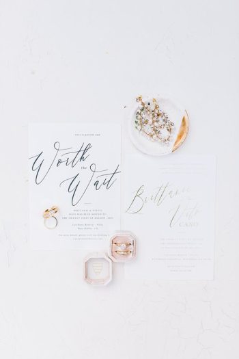 Elegant wedding invitation lay-flat with decorative elements and rings on a white background.