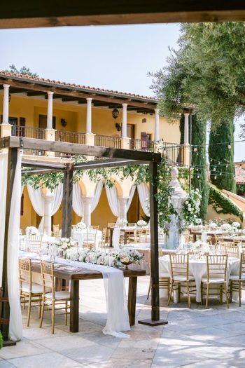 Elegant outdoor wedding reception setup with decorated tables under a pergola, adjacent to a building with a yellow facade.