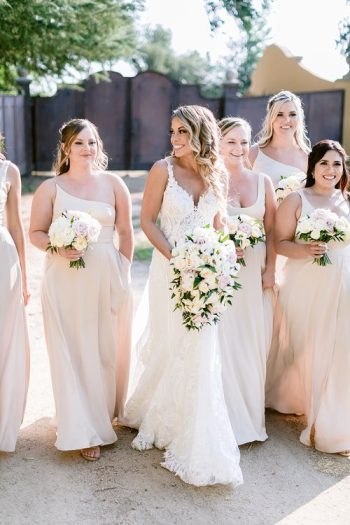 A bride in a white lace gown smiles while accompanied by her bridesmaids in matching blush dresses, all holding bouquets.
