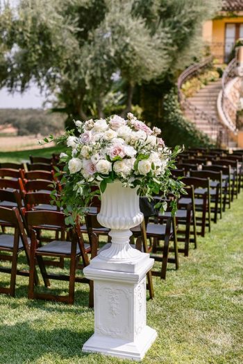 An elegant floral arrangement atop a white pedestal with rows of chairs set up for an outdoor event.