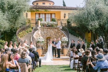 Outdoor wedding ceremony with a couple holding hands before an audience, set against a backdrop of a house with a balcony and surrounded by greenery.