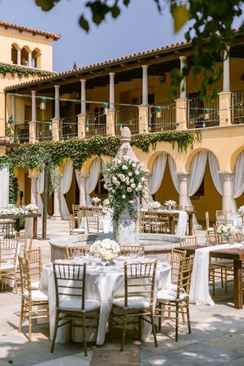 Elegant outdoor wedding reception setup with floral centerpieces and draped cabanas at a venue with mediterranean-style architecture.