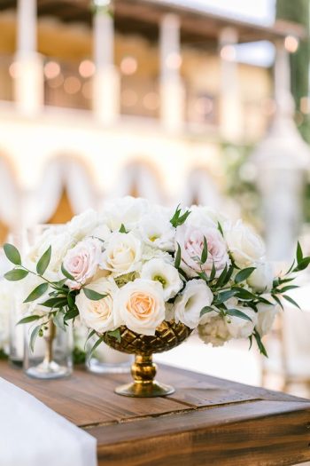 Elegant floral centerpiece with white and pastel roses in a gold vase on a wood table.