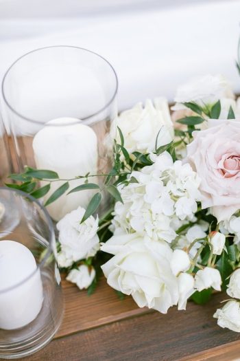 Elegant table centerpiece featuring white and blush roses, hydrangeas, greenery, and candles in tall glass holders.