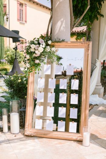 An ornate framed seating chart for a wedding event, adorned with flowers and flanked by candles.