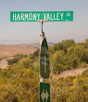 A road sign for harmony valley rd above a highway route marker for california state route 1, with a backdrop of hills and sparse vegetation.