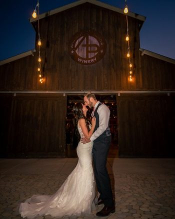 A couple sharing a kiss outside a building with the sign "winery," adorned with hanging lights against a twilight sky.