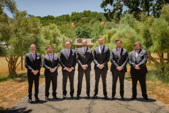 Group of seven men dressed in formal suits posing for a photograph outdoors.