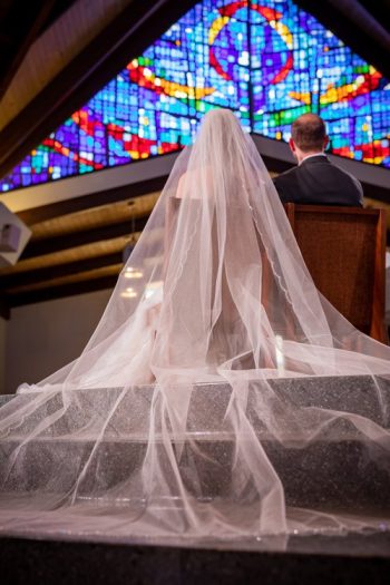 A bride walking up the aisle towards the groom in a church with a stained glass window in the background.