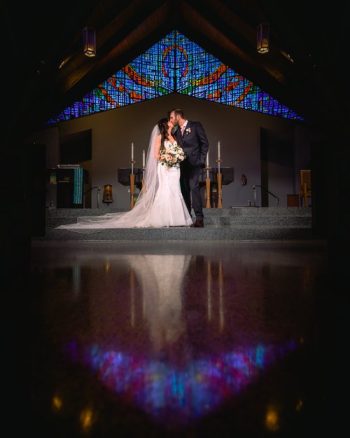A couple in wedding attire shares an intimate moment in front of a stained glass window in a church.