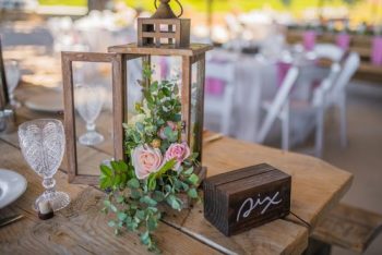 Elegant table setting with a floral arrangement inside a lantern, a wooden table number, and crystal stemware at a festive event.