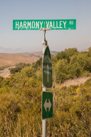 A road sign for harmony valley rd above a california hiking trail symbol, with a highway in the background.