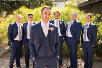 A group of six men in formal attire, with the foreground featuring a smiling groom and his groomsmen standing in the background.