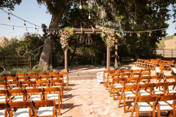 Outdoor wedding ceremony setup with rows of wooden chairs facing a floral-decorated arch and string lights.