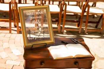 Guest book at an event displayed on a wooden podium with a sign inviting attendees to sign their names, with chairs arranged in the background.