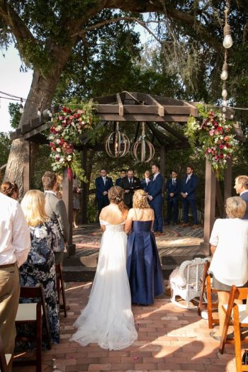 A wedding ceremony taking place outdoors under an arch decorated with flowers, with guests looking on.