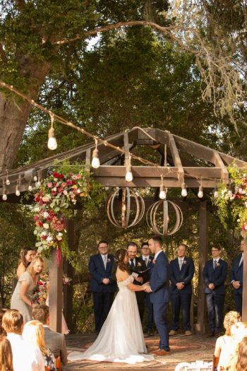 A couple exchanging vows at an outdoor wedding ceremony under a floral-decorated wooden arbor.