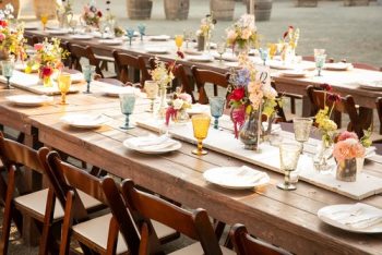 Outdoor dining setup with rustic wooden tables, assorted glassware, and colorful floral centerpieces.