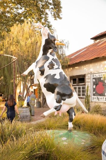 A large cow sculpture in a playful pose with visitors walking nearby at an outdoor venue with rustic buildings.