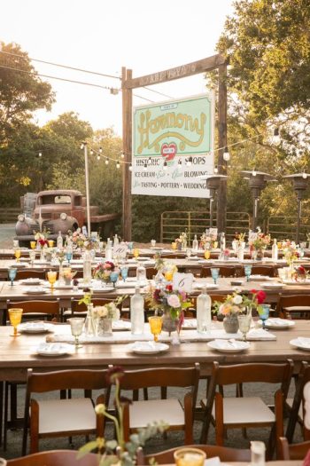 Outdoor rustic wedding reception setup with long tables, a vintage truck in the background, under warm evening light.