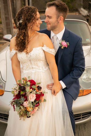 A bride and groom sharing a tender moment, with the bride holding a bouquet, and a vintage car in the background.
