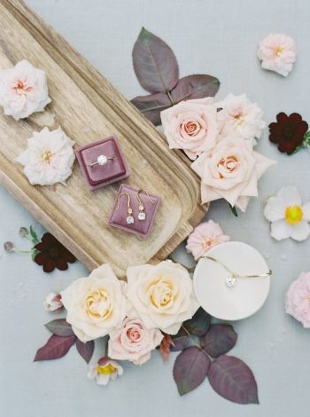 A jewelry presentation with a necklace and earrings arranged amongst pale roses and blossoms on a textured surface.