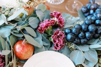 Elegant table setting with a floral and fruit centerpiece featuring dark grapes, a pomegranate, and red carnations amidst green foliage.