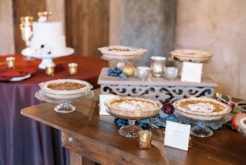An assortment of pies displayed on elegant stands at a dessert table with a wedding cake in the background.