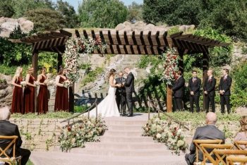 A wedding ceremony taking place outdoors with a bride and groom at the altar, flanked by bridesmaids in maroon dresses and groomsmen in black suits.