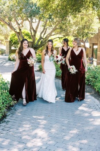 A bride and her bridesmaids walking and smiling on a pathway, each holding a bouquet, with the bridesmaids dressed in matching burgundy gowns.