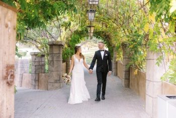 A bride and groom holding hands and walking under a vine-covered archway.